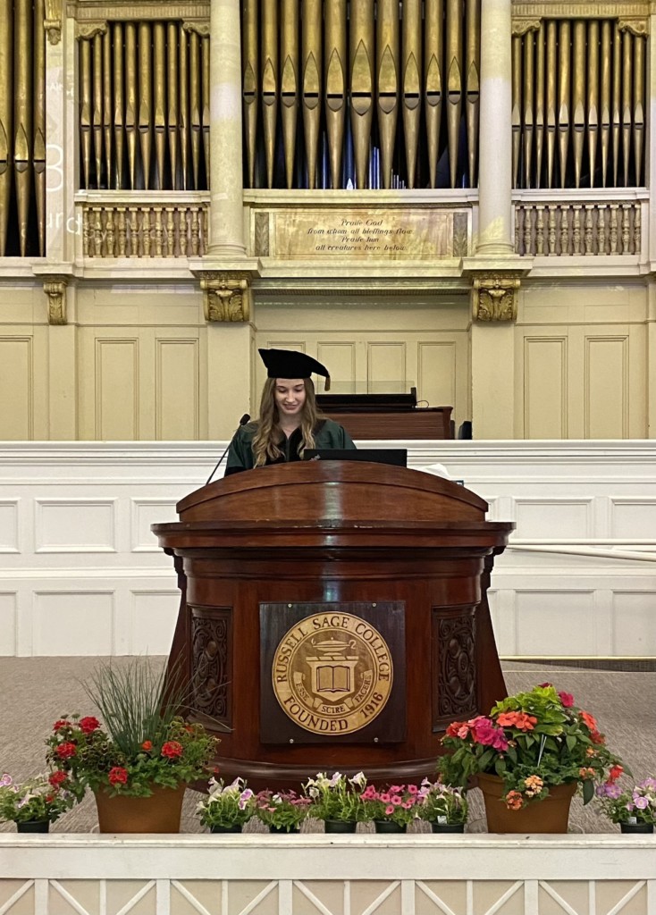 person in gradation cap and gown standing behind podium on a stage.